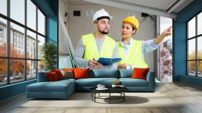 Caucasian man and asian woman builders discussing project documentation in apartment. Pointing finger gesture. Wall mural