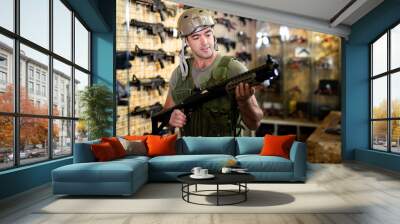 Caucasian man wearing armor vest and helmet while choosing machine gun in airsoft shop. Wall mural