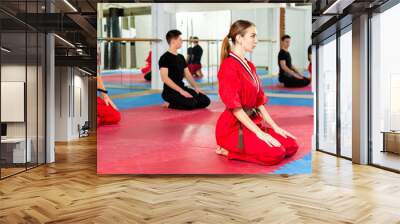 Concentrated young woman in red martial arts uniform sitting on her knees on mat in gym, ready for training with group Wall mural