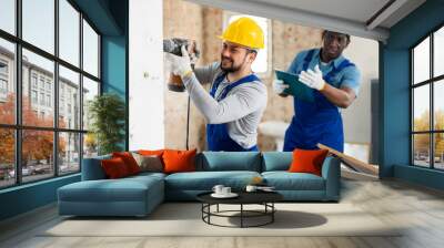 Confident man construction worker drilling wall with pneumatic hammer drill under control of foreman on indoor building construction site Wall mural