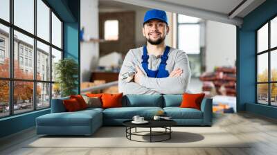 Confident positive young bearded builder wearing blue overalls and cap posing at construction site indoors.. Wall mural