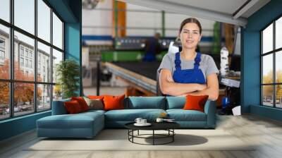 Confident smiling young female worker posing with crossed arms in glass factory, against blurred background of workshop setting, showcasing professionalism and pride in job Wall mural