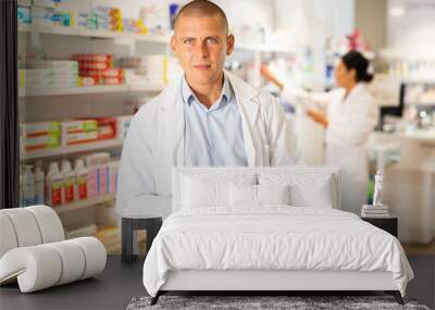 Doctor in white coat standing in salesroom with pan and recipe blanks in hands and looking in camera. His co-worker setting out drugs on shelves in background. Wall mural