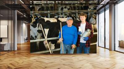 Elderly farmer and his assistant grandson with bottle of milk on the background of cows in a stall Wall mural