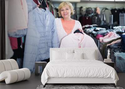 elderly positive blonde woman shopping and holding synthetic jackets in the fashion store Wall mural