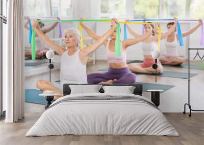 Elderly woman exercising with pilates elastic band on mat in female group in studio Wall mural