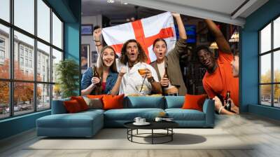 Excited young adult friends, sports fans celebrating victory of favorite football team with beer in bar, waving national flag of England Wall mural