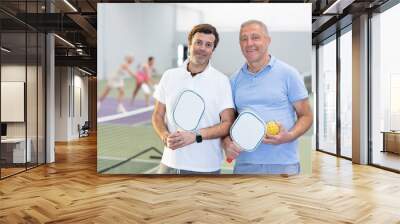 Experienced team of pickleball players, two friendly smiling men of different ages, standing on indoor court with paddles and balls in hand ready to training Wall mural