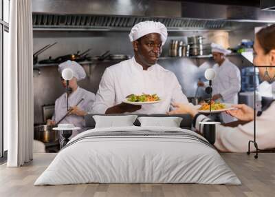 Focused young adult man chef giving out ready meals to waitress on order station in open restaurant kitchen Wall mural