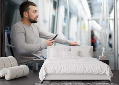 Focused young man traveling with suitcase in empty subway car, sitting on seat and browsing messages on his phone Wall mural