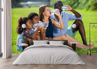 Friendly interracial family sitting on green lawn, relaxing and drinking water during family walk in city park on hot summer day.. Wall mural