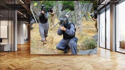 Group of men and women in protective clothes and helmets shooting with paintball markers. Teams fighting on battlefield outdoors. Wall mural