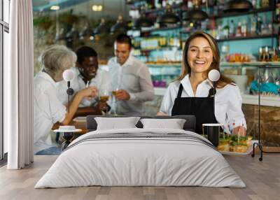 Happy Latin female waiter in uniform pointing hand on the plate with pinchos behind a table with diverse friends and bar counter Wall mural