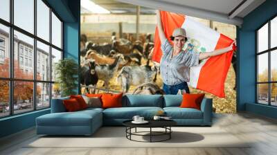 Happy young Latin female traveler in plaid shirt waving Peruvian flag during visit to livestock goat farm Wall mural