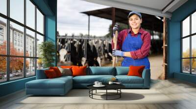 Hardworking young woman farmer in overalls posing with rake and smiling at camera while working on cow farm Wall mural