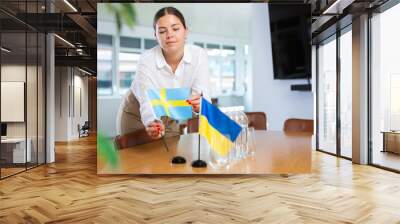 Interested young businesswoman setting national flags of Ukraine and Sweden on table in meeting room before negotiations. Concept of successful bilateral relations Wall mural