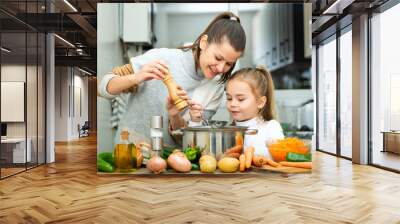 Little daughter helping cooking soup and mother add pepper to pan Wall mural