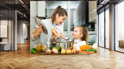 Little daughter helping cooking soup and mother add pepper to pan Wall mural