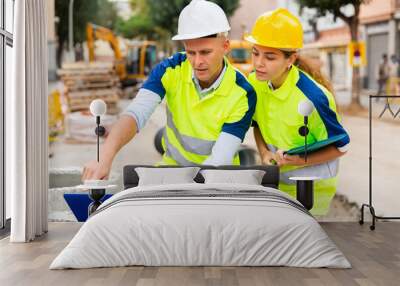 Man and woman builders in uniform and hardhats discussing while standing in construction site. Man using laptop, woman holding folder with documentation. Wall mural