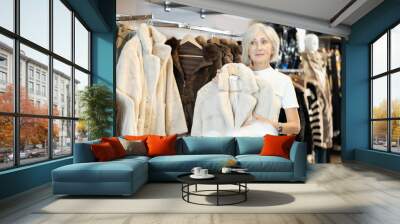 Mature lady carefully inspects fur coats, contemplating which one best complements her winter wardrobe. Wall mural