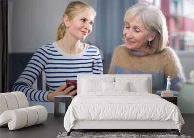 Mature woman with her adult daughter, sitting at a table in a room, looking at photos on a mobile phone Wall mural