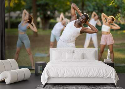 Middle-aged African American man in comfortable sportswear performing Tiryaka Tadasana, or Swaying Tree Pose, during group yoga session in green city park on summer day Wall mural