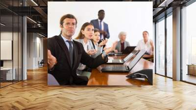 Middle aged white brunette male office worker with questioning facial expression spreading his arms to the sides as if asking what nonsense are you saying, while sitting at corporate team meeting in Wall mural