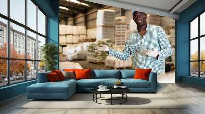 Portrait of african-american man, construction material storage worker, standing inside warehouse and making presenting gesture. Wall mural