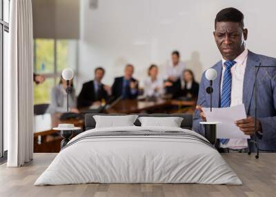 Portrait of african-american office worker in suit failed his report. Upset company worker holding papers, standing in meeting room during conference. Wall mural