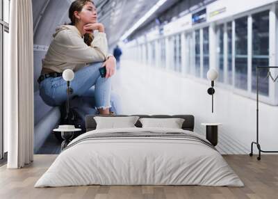 Portrait of thoughtful girl sitting on a bench at a metro station waiting for a train, holding a mobile phone in her hand Wall mural