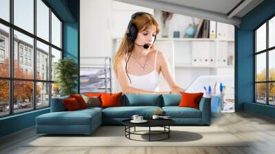 Portrait of young woman wearing headset, using laptop. Call center worker sitting at desk in office. Wall mural