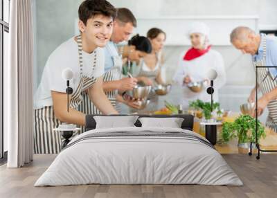 Positive interested young guy attending group culinary classes, standing by table with ingredients and utensils, learning culinary skills from professional chef Wall mural