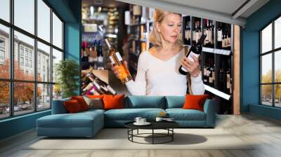 Positive middle aged blonde female choosing bottled wine in modern wineshop Wall mural