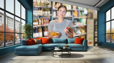 Positive thoughtful young female customer comparing two bottles of juice while shopping for organic beverages in local grocery store Wall mural