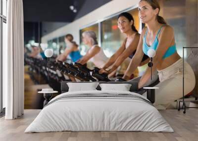 Positive young european female riding exercise bike during cycling class in gym Wall mural