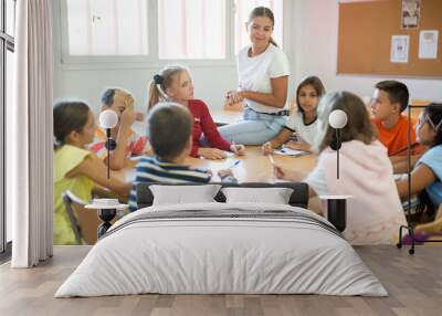 Positive young female teacher giving lesson for group of preteen schoolchildren sitting around common desk and listening attentively Wall mural