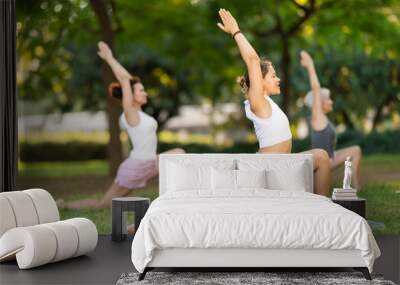 Positive young girl doing yoga on mat during group outdoor class in green sunlit summer park, performing Anjaneyasana or Crescent Low Lunge Pose, demonstrating flexibility and balance Wall mural