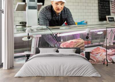 Smiling experienced adult butcher working behind counter in butchery, offering piece of fresh raw beef chop on ribs Wall mural