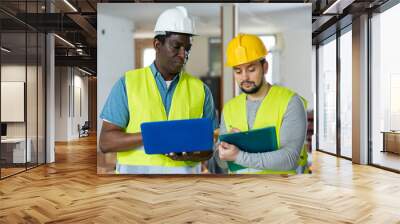 Two skilled maintenance workers wearing protective helmets and vests talking work progress and gesturing at indoor building site Wall mural