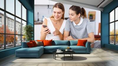 Two young girls having conversation and looking at smartphone in kitchen Wall mural