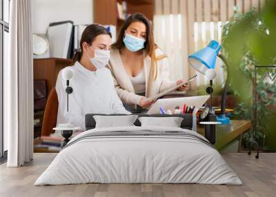 Woman assistant in face mask standing with documents near her female boss in office Wall mural