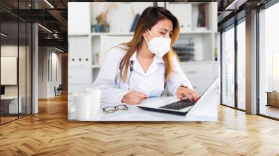 Woman doctor in medical mask is working at laptop in clinic Wall mural