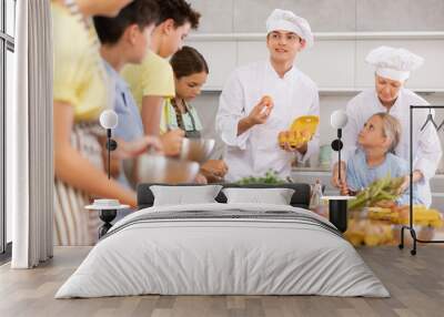 Young guy and adult woman cook in uniform teaches group of children how to cook dish Wall mural