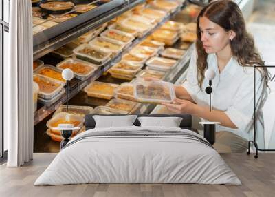 Young positive woman choosing ready meal in plastic container at supermarket Wall mural