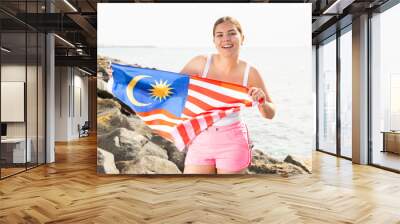 Young smiling woman in summer clothes posing with big flag of Malaysia on sea coast Wall mural