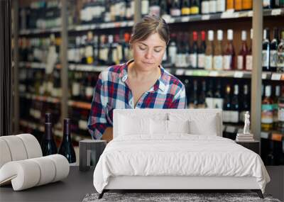 Young woman carefully reads the label on bottle while choosing wine in a supermarket Wall mural