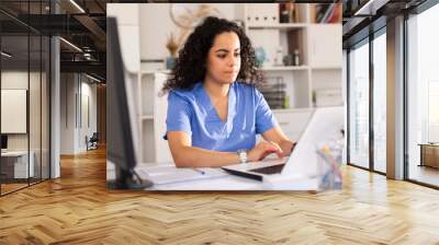 Young woman doctor assistant working in medical office using laptop computer and writing prescription Wall mural