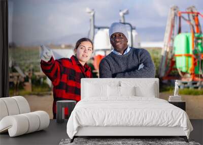 Young woman farmer, standing against the background of an agricultural machine, discusses important work issues with an ..african american man colleague, shows him something, pointing to it Wall mural