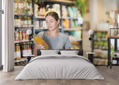 Young woman in bio supermarket buys drink. Girl buyer examines packaging bottle of product and reads information on label Wall mural