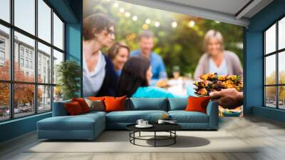  group of friends in their forties gathered around a table in the garden to share a bbq meal. A man offers chicken skewers to guests Wall mural